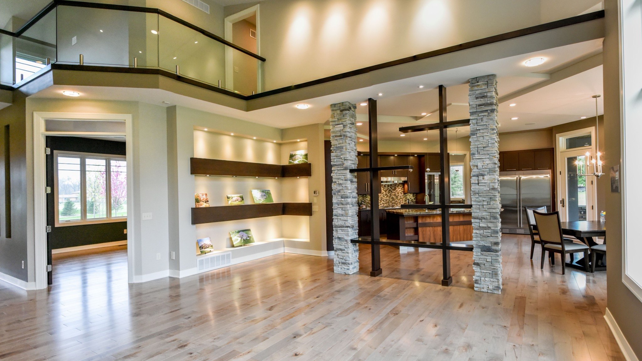 Open custom home interior with stone columns, kitchen, and glass-railed upper level